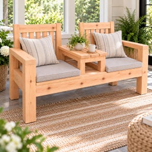 May include: A light-colored wooden bench featuring two seats separated by a central table. The bench includes two light gray seat cushions, two striped throw pillows, and a small potted green plant and a white mug on the center table.
