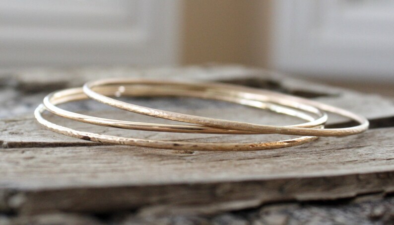 May include: Three gold bangles stacked on top of each other. The bangles are hammered and have a textured surface.
