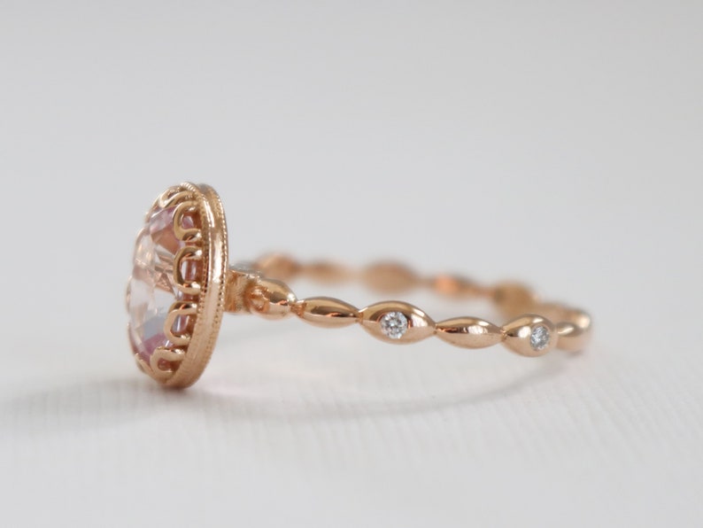 May include: A rose gold ring with a large oval-shaped pink gemstone set in a delicate filigree design. The band is adorned with small round diamonds set in a scalloped pattern.