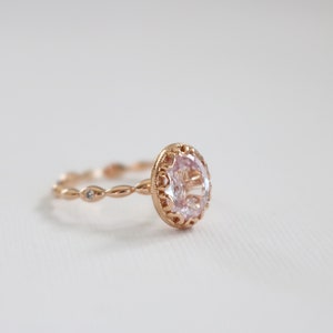 May include: A delicate rose gold ring with a large oval-shaped clear gemstone set in a detailed filigree design. The band is adorned with smaller clear gemstones.