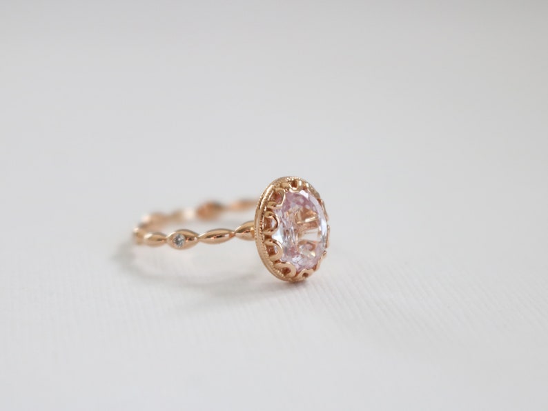 May include: A delicate rose gold ring with a large oval-shaped clear gemstone set in a detailed filigree design. The band is adorned with smaller clear gemstones.
