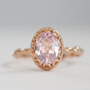 May include: A delicate rose gold ring with a pink oval gemstone set in a detailed filigree design. The band is adorned with smaller gemstones.