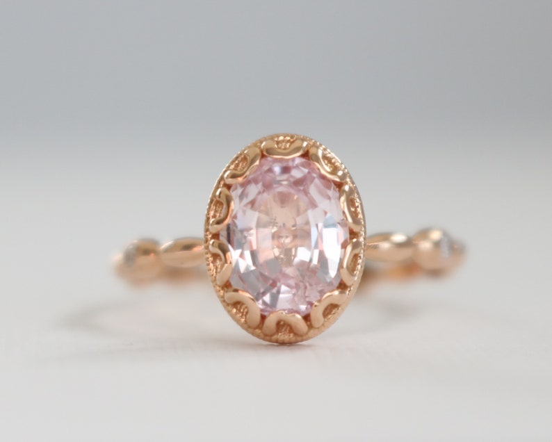 May include: A delicate rose gold ring with a pink oval gemstone set in a detailed filigree design. The band is adorned with smaller gemstones.