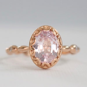 May include: A delicate rose gold ring with a pink gemstone set in a detailed filigree design. The band is adorned with small diamonds.