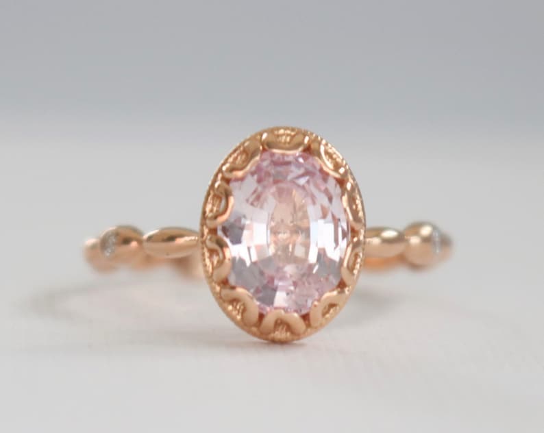 May include: A delicate rose gold ring with a pink gemstone set in a detailed filigree design. The band is adorned with small diamonds.