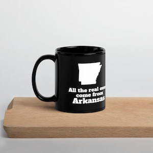 May include: A black ceramic mug featuring a white silhouette of the state of Arkansas. Below the state shape, the text reads, "All the real ones come from Arkansas" in a bold, white, sans-serif font. The mug has a curved handle on the left side.