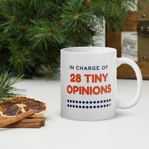 Peut inclure: Un mug en céramique blanche avec le texte bleu foncé IN CHARGE OF et le texte orange 28 TINY OPINIONS, suivi de deux rangées de points bleu foncé avec un seul point orange à la fin de la rangée inférieure, imprimés sur le devant du mug.