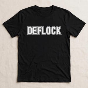 May include: A black short-sleeved t-shirt featuring the word DEFLOCK printed in bold white capital letters with a thin black outline across the center of the chest. The garment has a crew neckline and a standard fit with short sleeves.