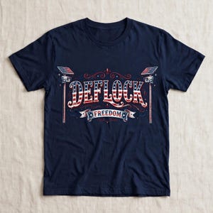 Peut inclure: Un t-shirt bleu marine à manches courtes avec une impression graphique portant le texte DEFLOCK et FREEDOM. Les lettres sont remplies d'un motif de drapeau américain rouge, blanc et bleu, flanquées de deux caméras de surveillance sur poteaux.