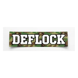 May include: A rectangular sticker featuring a camouflage pattern in shades of green, brown, and black. The word DEFLOCK is printed across the center in bold, white, block-style capital letters with a thick black outline.