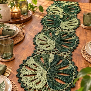 May include: A long, rectangular crochet table runner featuring a repeating pattern of circular monstera leaf motifs in varying shades of dark and light green. The runner has a scalloped, intricate border and is crafted from yarn with a textured finish.