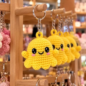 May include: A bright yellow crocheted octopus keychain featuring a smiling face with large black eyes and small pink cheeks. The plush octopus is attached to a silver metal keyring and chain, hanging alongside other crocheted flower keychains.