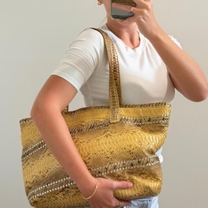 May include: A large yellow tote bag featuring a textured snakeskin pattern with horizontal bands of metallic silver studs. The bag has two matching shoulder straps and a rectangular shape, showcasing a detailed scale-like surface texture throughout.
