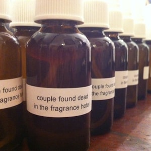 May include: A row of brown glass bottles with white caps and labels. The labels read "couple found dead in the fragrance hotel". The bottles are filled with a dark liquid.