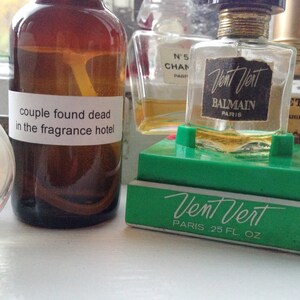 May include: A collection of perfume bottles, including a brown bottle with a label that reads "couple found dead in the fragrance hotel." A "Vent Vert" perfume bottle is displayed on a green stand.
