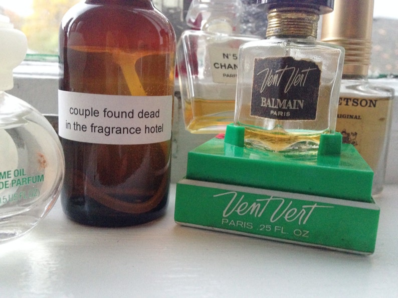 May include: A collection of perfume bottles, including a brown bottle with a label that reads "couple found dead in the fragrance hotel." A "Vent Vert" perfume bottle is displayed on a green stand.