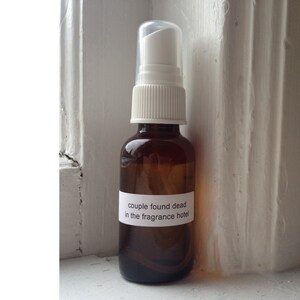 May include: A small, brown glass spray bottle with a white pump and cap. A white label on the bottle reads "couple found dead in the fragrance hotel."