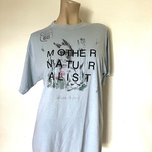 May include: A light blue t-shirt with the text "MOTHER NATURALIST" in black block letters. The shirt features a graphic of a rabbit, cacti, and flowers. The Route 66 logo is printed on the top left.