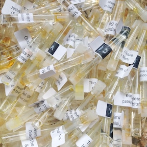 May include: A collection of small, clear glass vials filled with a light amber liquid. Each vial has a white label with black text. The vials are scattered, creating a textured, close-up view. Some labels are partially visible.
