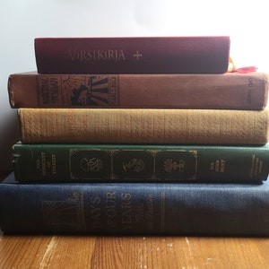 May include: A stack of five books with different colored covers. The top book is red with the text "VIRSIKIRJA +" on the cover. The second book is brown with the text "MATERIAL WOMAN" on the cover. The third book is beige with a textured cover. The fourth book is green with the text "THE CONQUEST OF EVEREST" on the cover. The bottom book is blue with the text "DAYS OF OUR YEARS" on the cover.
