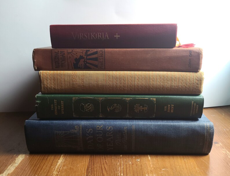 May include: A stack of five books with different colored covers. The top book is red with the text "VIRSIKIRJA +" on the cover. The second book is brown with the text "MATERIAL WOMAN" on the cover. The third book is beige with a textured cover. The fourth book is green with the text "THE CONQUEST OF EVEREST" on the cover. The bottom book is blue with the text "DAYS OF OUR YEARS" on the cover.