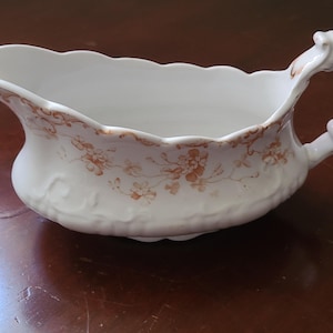May include: White porcelain gravy boat with a scalloped rim and a decorative handle. The boat features a floral pattern in shades of brown and has a textured surface. A classic piece for serving sauces and gravies.