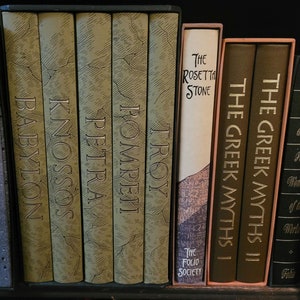 May include: A bookshelf with a collection of hardcover books with gold lettering. The books are arranged in a row and include titles like "Troy", "Pompeii", "Petra", "Knossos", "Babylon", "The Rosetta Stone", "The Greek Myths I", "The Greek Myths II", and "What are the Seven Wonders of the World?"