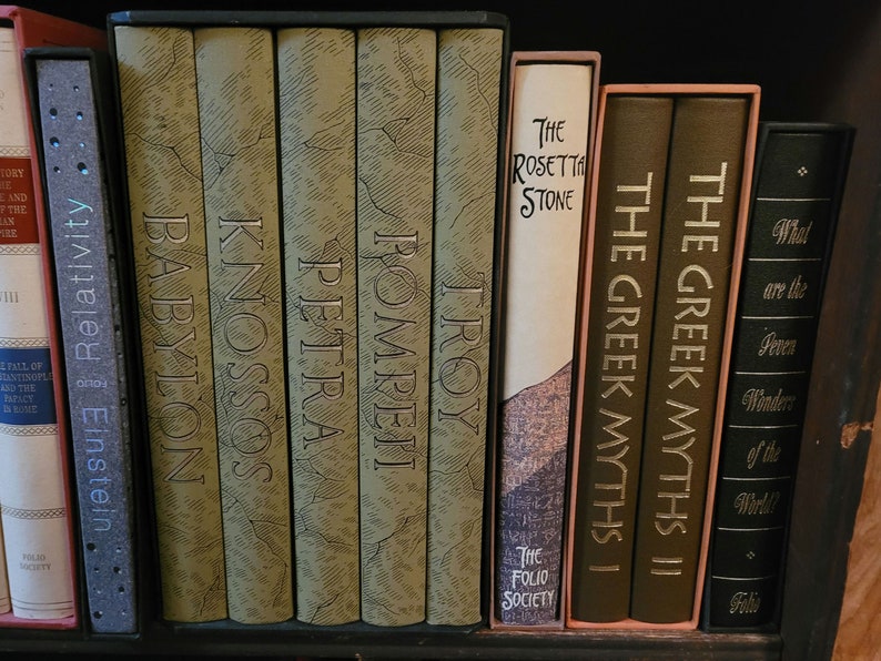 May include: A bookshelf with a collection of hardcover books with gold lettering. The books are arranged in a row and include titles like "Troy", "Pompeii", "Petra", "Knossos", "Babylon", "The Rosetta Stone", "The Greek Myths I", "The Greek Myths II", and "What are the Seven Wonders of the World?"