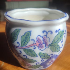 May include: A small white ceramic bowl with a scalloped rim and a blue painted edge. The exterior features a repeating pattern of green leaves, pink flowers, and blue blossoms. The bowl has a rounded shape and a smooth, glossy finish throughout.