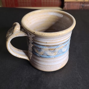 May include: Handmade ceramic mug with a unique handle design. The mug features a light beige and blue wavy pattern around the middle, with a light beige interior and rim. The mug is perfect for coffee, tea, or any hot beverage.