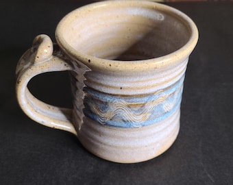 May include: Handmade ceramic mug with a unique handle design. The mug features a light beige and blue wavy pattern around the middle, with a light beige interior and rim. The mug is perfect for coffee, tea, or any hot beverage.