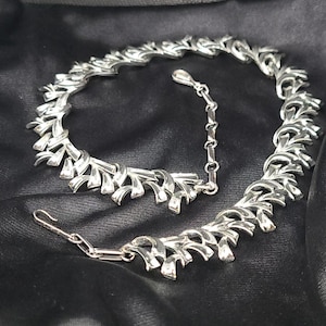 May include: A silver tone necklace with a geometric design. The necklace is made of interlocking pieces that resemble leaves. The necklace has a clasp closure.