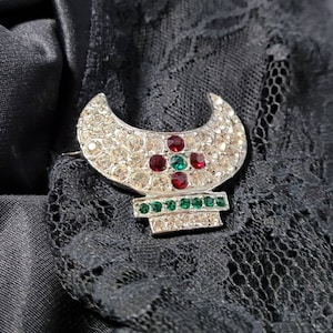 May include: A silver-toned brooch with a crescent-shaped top, encrusted with clear rhinestones. The center features a cross design with red and green stones. The bottom section has a row of green stones. The brooch is set against a black lace background.