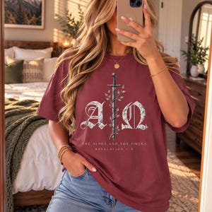 May include: A maroon short-sleeve t-shirt featuring a graphic of a sword entwined with vines between the letters A and Ω. Below the graphic, the text reads THE ALPHA AND THE OMEGA REVELATION 1:8 in white capital letters.