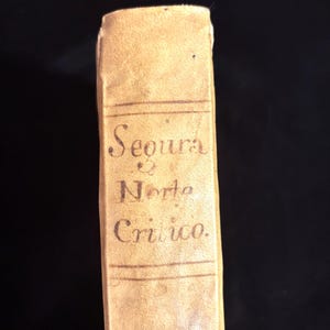 May include: The spine of a tan, aged book featuring the printed text Segura Norta Critico. The spine includes horizontal lines above and below the title, with a worn, textured surface and visible signs of age on the paper cover.