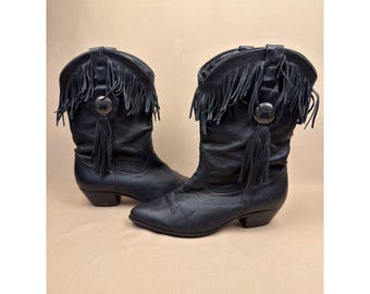 May include: A pair of black mid-calf boots featuring fringe detailing around the upper shaft, a pointed toe, a low stacked heel, and a circular silver-toned metal concho ornament with a hanging tassel on the outer side of each boot.