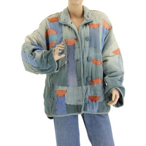 Könnte beinhalten: Eine blaue gesteppte Jacke mit Patchwork-Design, das geometrische Formen in Blautönen, Orange und Rot zeigt. Die Jacke hat einen Knopfverschluss und einen Kragen.