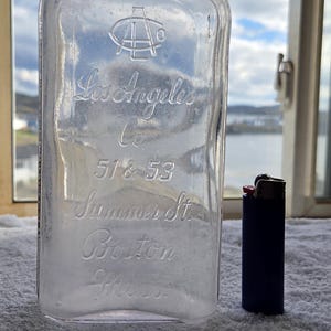 May include: A clear glass bottle featuring an embossed logo and text reading Los Angeles Co 51 & 53 Summer St Boston Mass. The bottle is placed beside a dark blue rectangular lighter for scale, illustrating the relative height of the glass vessel.