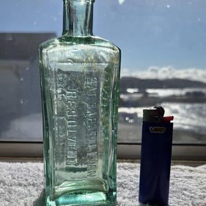 May include: A clear, light green glass bottle with a rectangular body and a narrow neck. Embossed text on the front reads "SARSAPARILLIAN RESOLVENT". A blue Bic lighter is positioned next to the bottle for scale, showing the relative size.