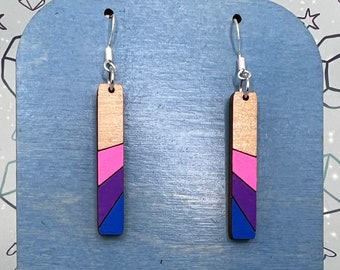 Könnte beinhalten: Ein Paar Hängeohrringe mit einem Holzstab, der in rosa, blauen und violetten Streifen bemalt ist. Die Ohrringe werden auf einem blauen Holzständer mit dem Text "Souvrin's Art" auf der Unterseite präsentiert.