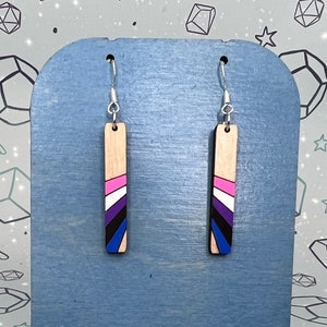 May include: A pair of wooden earrings with a blue, pink, white and black striped design. The earrings are hanging on a blue display stand with the text "Sourin's Art" printed on it.