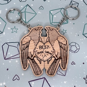 May include: Two wooden keychains shaped like birds facing each other. The birds are holding a heart-shaped piece of wood with the text 'Best Trash Goblins' on it. The keychains are attached to silver keyrings.