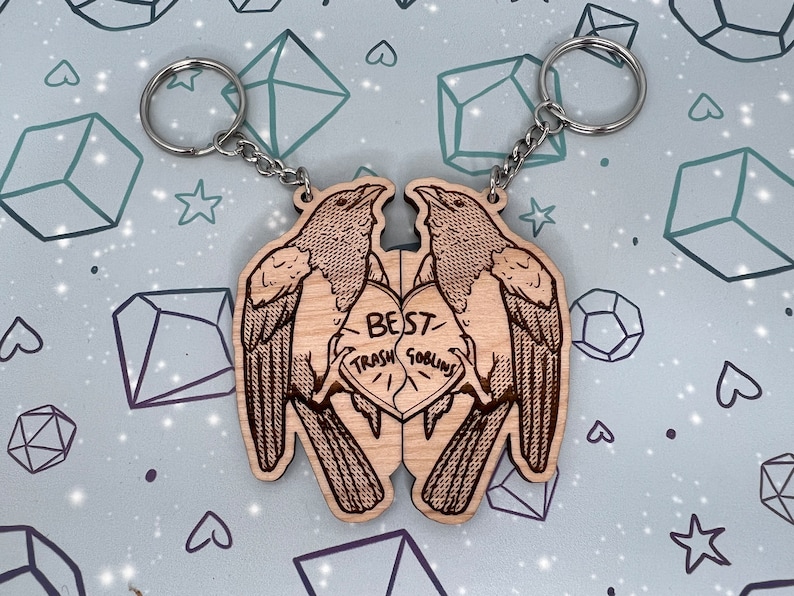 May include: Two wooden keychains shaped like birds facing each other. The birds are holding a heart-shaped piece of wood with the text 'Best Trash Goblins' on it. The keychains are attached to silver keyrings.