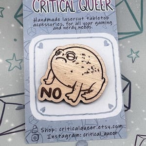Puede incluir: Un pin de madera con forma de rana con la palabra "NO" en una tarjeta. La tarjeta tiene el texto "CRITICAL QUEER" y "Accesorios de mesa cortados con láser hechos a mano, para todas tus necesidades de juego y nerd".