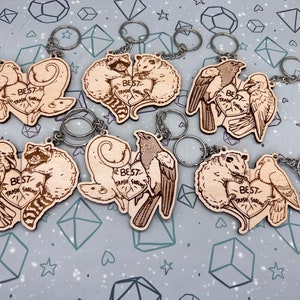 May include: Six wooden heart-shaped keychains with different animal pairs. Each keychain has a silver key ring and features a pair of animals with the text 'Best Trash Friends' in a heart shape. The animals include a raccoon and a possum, a magpie and a possum, a pair of crows, a pair of pigeons, and a raccoon and a possum.