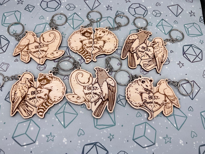 May include: Six wooden heart-shaped keychains with different animal pairs. Each keychain has a silver key ring and features a pair of animals with the text 'Best Trash Friends' in a heart shape. The animals include a raccoon and a possum, a magpie and a possum, a pair of crows, a pair of pigeons, and a raccoon and a possum.