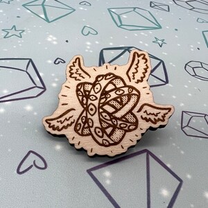 May include: A wooden pin with a detailed design of a winged eye. The eye is surrounded by a swirling pattern and has a halo around it.