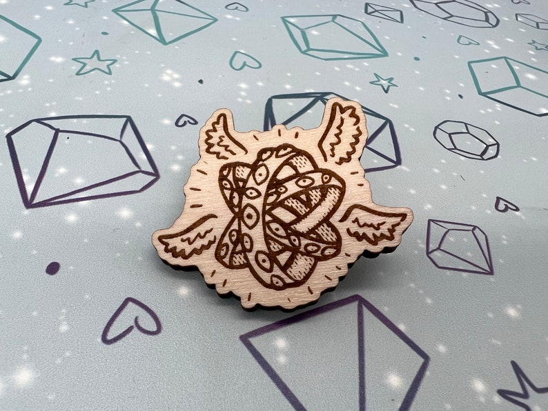 May include: A wooden pin with a detailed design of a winged eye. The eye is surrounded by a swirling pattern and has a halo around it.