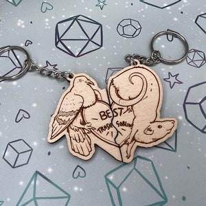 May include: Two wooden keychains shaped like a dove and a rat. The dove is on the left and the rat is on the right. The keychains are connected by a heart-shaped piece with the text 'BEST TRASH GOBLIN' written on it. The keychains are attached to a silver chain with a silver key ring.