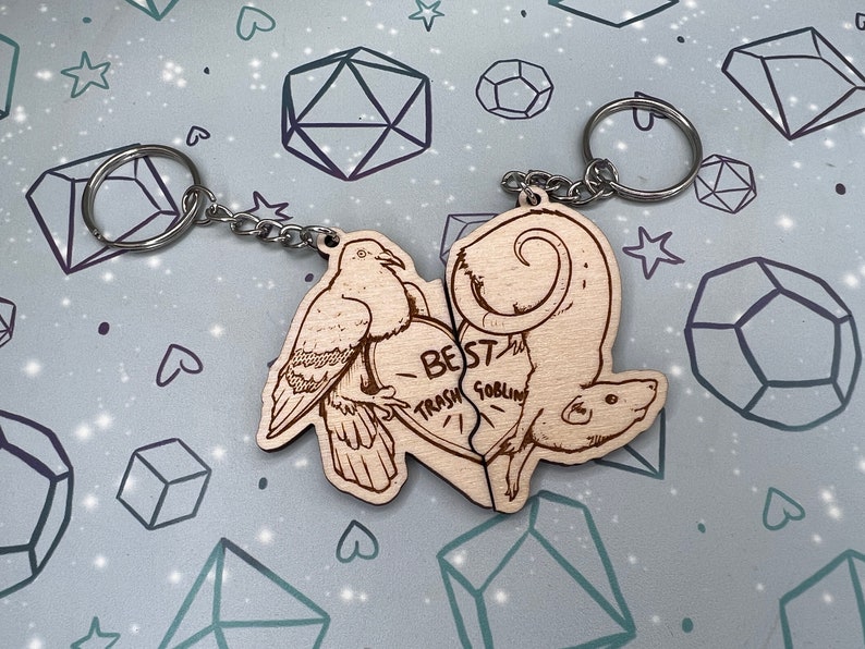 May include: Two wooden keychains shaped like a dove and a rat. The dove is on the left and the rat is on the right. The keychains are connected by a heart-shaped piece with the text 'BEST TRASH GOBLIN' written on it. The keychains are attached to a silver chain with a silver key ring.